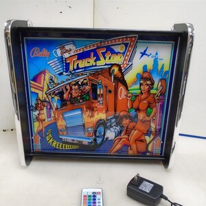 May include: A black framed light-up sign with a chrome border. The sign features a colourful illustration of a semi-truck with the words "Truck Stop" in a red and white banner. The sign is plugged in and has a remote control with coloured buttons.