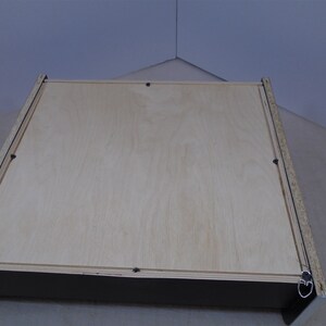 May include: The back of a wooden picture frame with a light wood finish. The frame has a black border and two metal hanging hooks.