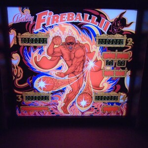 May include: A pinball machine with a red, orange, and blue background. The game is called "Fireball II" and features a muscular figure with a cape and a devil's head in the upper right corner. The game has multiple score displays.