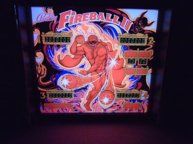May include: A pinball machine with a red, orange, and blue background. The game is called "Fireball II" and features a muscular figure with a cape and a devil's head in the upper right corner. The game has multiple score displays.