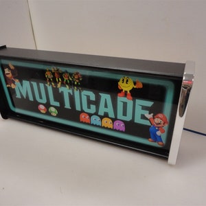 May include: A black and silver sign with the word "MULTICADE" in white letters on a teal background. The sign features various video game characters, including Mario, Pac-Man, Donkey Kong, and the Teenage Mutant Ninja Turtles.