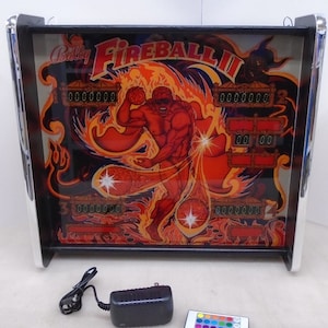 May include: A miniature pinball machine with a black cabinet and a glass playfield featuring a red, orange, and yellow illustration of a muscular figure with the text "Bally Fireball II".