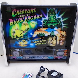 Puede incluir: Una caja de luz enmarcada con el cartel de la película "Creature from the Black Lagoon". El arte del cartel incluye la criatura, una mujer y el texto "Creature from the Black Lagoon 3-D". El marco tiene un borde negro y ribete plateado.