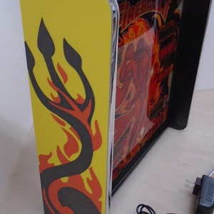 May include: A yellow and black pinball machine with a red and orange flame design. The machine is lit up and has a black frame. The machine is plugged in and has a power cord.