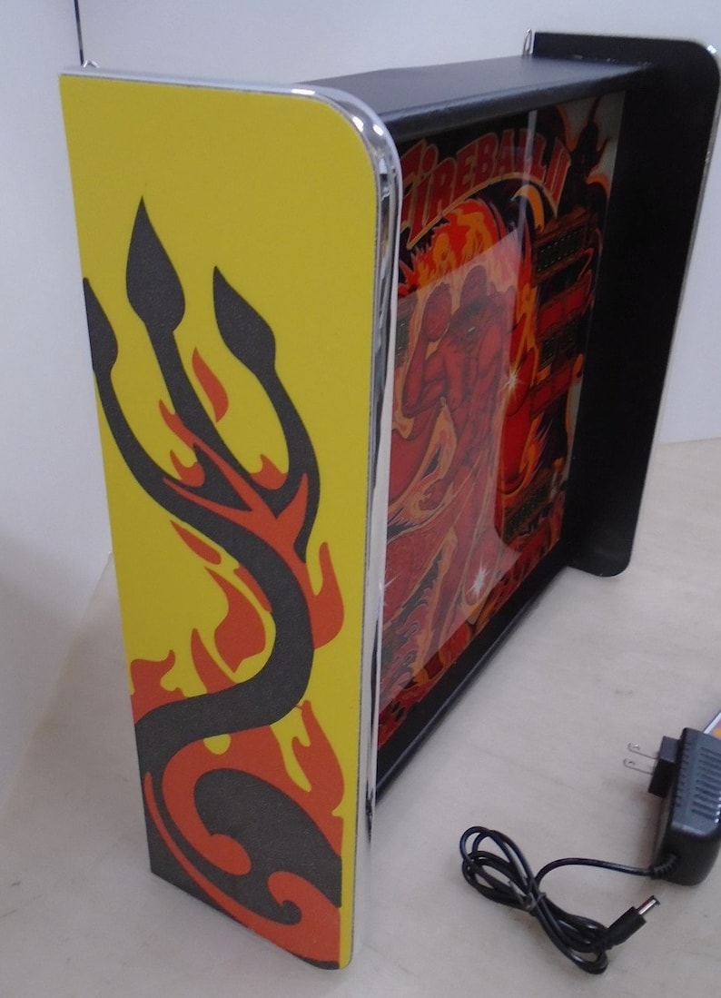 May include: A yellow and black pinball machine with a red and orange flame design. The machine is lit up and has a black frame. The machine is plugged in and has a power cord.