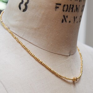 May include: A delicate gold-colored necklace with small, round beads strung together. The necklace is displayed on a mannequin. The necklace has a small, round clasp. The necklace is a jewelry item.