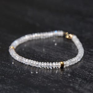 May include: A delicate bracelet featuring small, round, translucent beads. The beads are strung together, forming a circular shape. Gold-colored metal accents are visible at the clasp and at intervals along the bracelet. The bracelet is displayed on a dark surface.