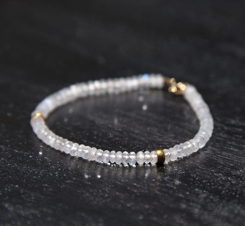May include: A delicate bracelet featuring small, round, translucent beads. The beads are strung together, forming a circular shape. Gold-colored metal accents are visible at the clasp and at intervals along the bracelet. The bracelet is displayed on a dark surface.