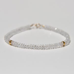 May include: A delicate bracelet featuring small, clear beads strung together. The bracelet has two small gold-colored beads and a silver clasp. The jewelry is displayed on a white surface.