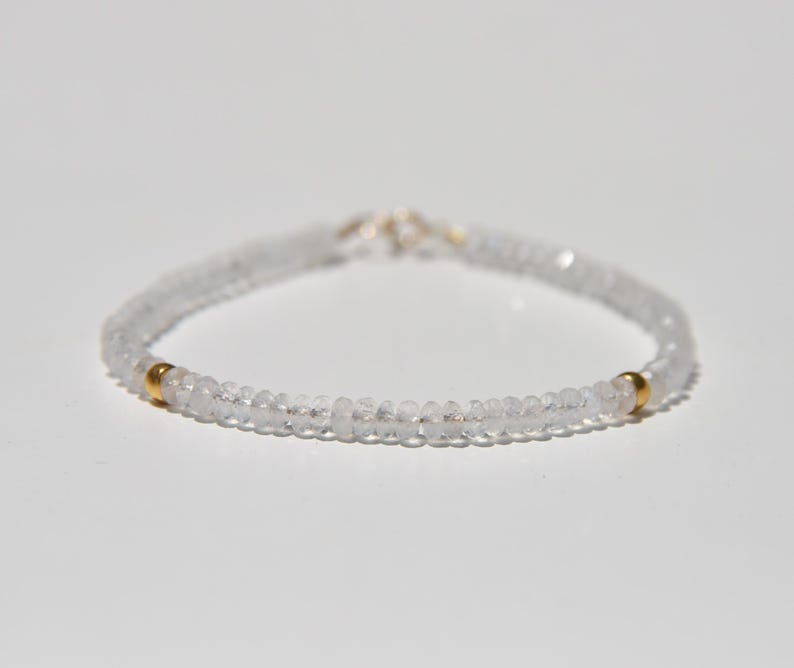 May include: A delicate bracelet featuring small, clear beads strung together. The bracelet has two small gold-colored beads and a silver clasp. The jewelry is displayed on a white surface.