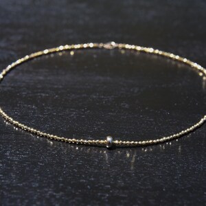 May include: Delicate gold-colored necklace with a beaded design. The necklace features a small, silver-colored bead near the center. The necklace is displayed on a dark surface.