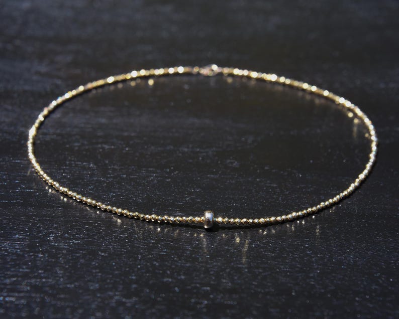 May include: Delicate gold-colored necklace with a beaded design. The necklace features a small, silver-colored bead near the center. The necklace is displayed on a dark surface.