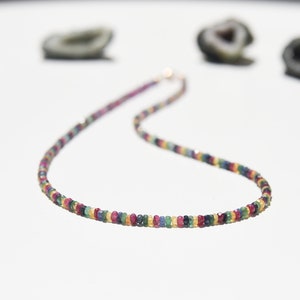 Pu&ograve; includere: Una collana colorata con piccole gemme rotonde in varie tonalit&agrave; di rosso, blu, giallo e verde. La collana ha una delicata chiusura dorata ed &egrave; esposta su una superficie bianca.