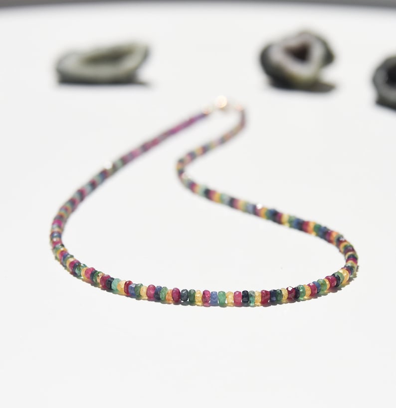 Pu&ograve; includere: Una collana colorata con piccole gemme rotonde in varie tonalit&agrave; di rosso, blu, giallo e verde. La collana ha una delicata chiusura dorata ed &egrave; esposta su una superficie bianca.
