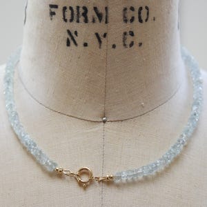 May include: A necklace featuring aquamarine gemstones and a gold clasp. The pale blue beads are strung together, forming an elegant piece of jewellery. The necklace is displayed on a mannequin.