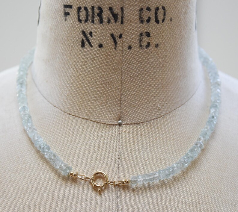 May include: A necklace featuring aquamarine gemstones and a gold clasp. The pale blue beads are strung together, forming an elegant piece of jewellery. The necklace is displayed on a mannequin.