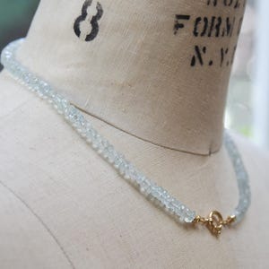 May include: A delicate aquamarine gemstone necklace with a gold clasp. The necklace is made of small, light blue-green beads. The clasp is a simple, round design. The necklace is displayed on a mannequin.