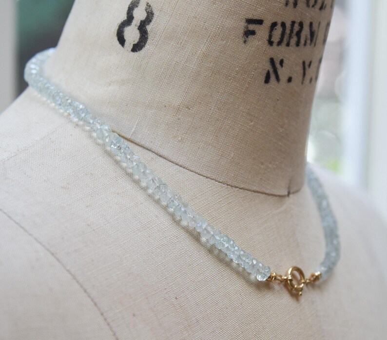 May include: A delicate aquamarine gemstone necklace with a gold clasp. The necklace is made of small, light blue-green beads. The clasp is a simple, round design. The necklace is displayed on a mannequin.