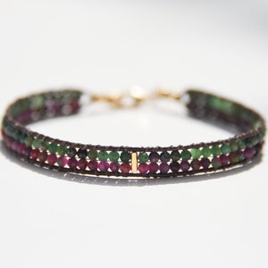 May include: A multi-strand bracelet with green and pink gemstones and a gold accent. The bracelet is made with brown leather cord.