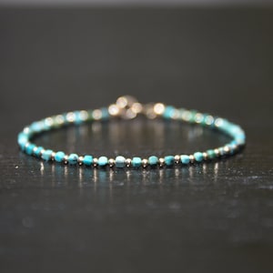 May include: A delicate bracelet featuring turquoise and silver beads. Small, square turquoise beads are interspersed with tiny silver beads. The clasp is silver. The bracelet is shown on a dark surface.