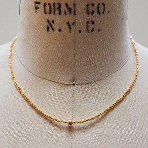 May include: A delicate gold necklace with small, faceted beads and a single, larger bead pendant. The necklace is displayed on a mannequin, showcasing its simple elegance and design.