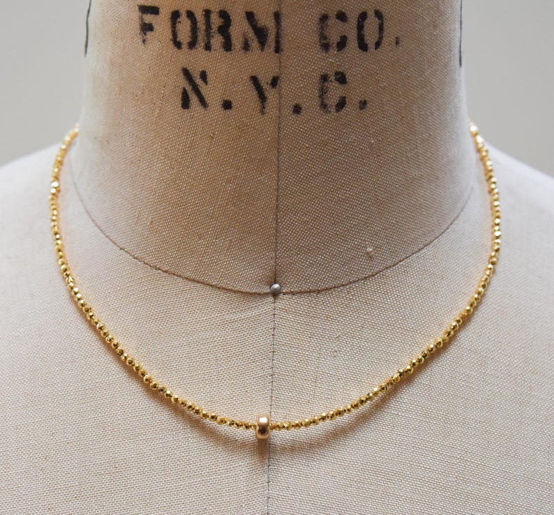 May include: A delicate gold necklace with small, faceted beads and a single, larger bead pendant. The necklace is displayed on a mannequin, showcasing its simple elegance and design.