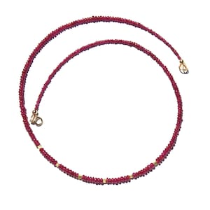 May include: A delicate necklace composed of small, faceted deep red ruby beads strung together. The strand is accented with tiny, polished gold-toned metal spacers at intervals and features a gold-toned metal clasp mechanism at the ends.