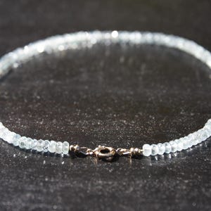 May include: A delicate necklace made of small, light blue gemstones. The necklace is fastened with a silver-coloured clasp. The gemstones, likely aquamarine, are strung together. The necklace is displayed on a dark surface.