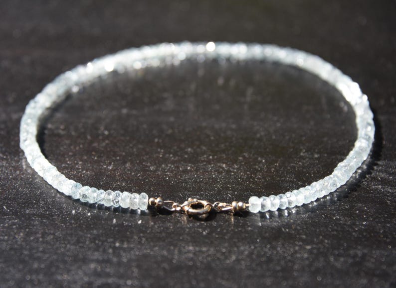 May include: A delicate necklace made of small, light blue gemstones. The necklace is fastened with a silver-coloured clasp. The gemstones, likely aquamarine, are strung together. The necklace is displayed on a dark surface.