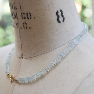May include: A light blue aquamarine necklace on a mannequin. The necklace is made of small, pale blue gemstones. It is secured with a gold-coloured clasp. The mannequin is a neutral beige colour, and the background is blurred.