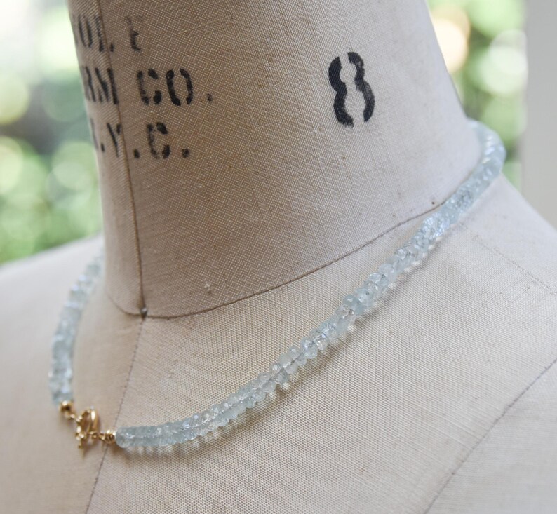 May include: A light blue aquamarine necklace on a mannequin. The necklace is made of small, pale blue gemstones. It is secured with a gold-coloured clasp. The mannequin is a neutral beige colour, and the background is blurred.