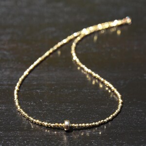 May include: A delicate gold necklace with a beaded chain and a small, silver-colored cylindrical accent. The necklace is displayed on a dark surface, highlighting its subtle shine and intricate design. The necklace is a fine piece of jewelry.