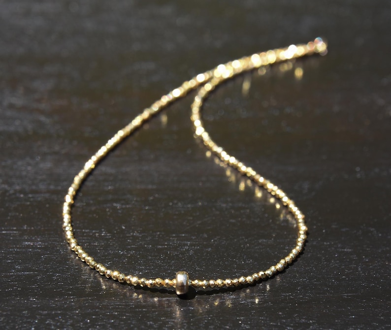 May include: A delicate gold necklace with a beaded chain and a small, silver-colored cylindrical accent. The necklace is displayed on a dark surface, highlighting its subtle shine and intricate design. The necklace is a fine piece of jewelry.