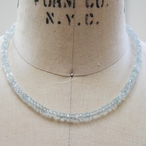 May include: A delicate aquamarine necklace made of small, faceted beads. The light blue gemstones are strung together, forming a choker-style necklace. The necklace is displayed on a vintage dress form.