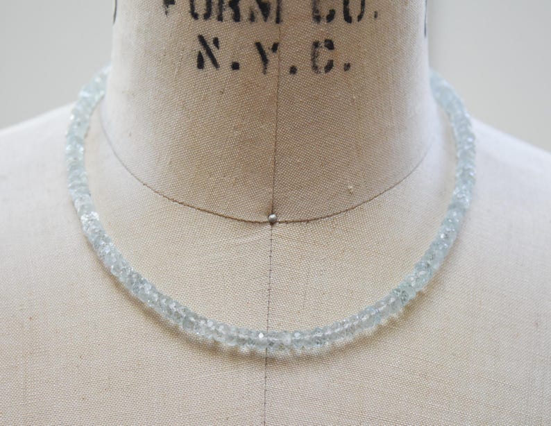 May include: A delicate aquamarine necklace made of small, faceted beads. The light blue gemstones are strung together, forming a choker-style necklace. The necklace is displayed on a vintage dress form.
