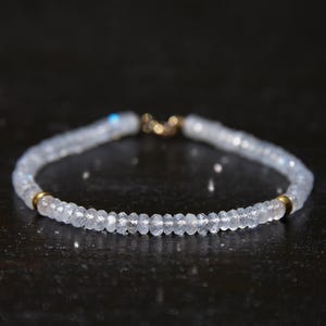 May include: A delicate bracelet featuring small, faceted, light blue beads strung together. The bracelet has gold-colored accents at the clasp and near the center. The beads appear to be semi-translucent, reflecting light.