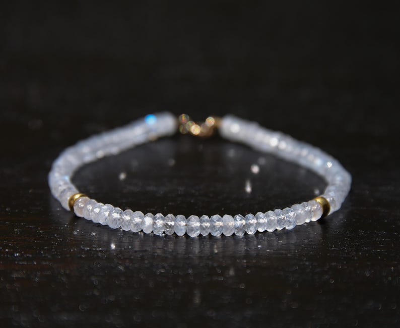 May include: A delicate bracelet featuring small, faceted, light blue beads strung together. The bracelet has gold-colored accents at the clasp and near the center. The beads appear to be semi-translucent, reflecting light.
