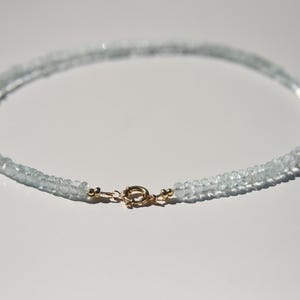 May include: A delicate aquamarine necklace with a gold clasp. The necklace is made of small, light blue gemstones, strung together to form a double strand. The clasp is a small, round design with a gold finish.