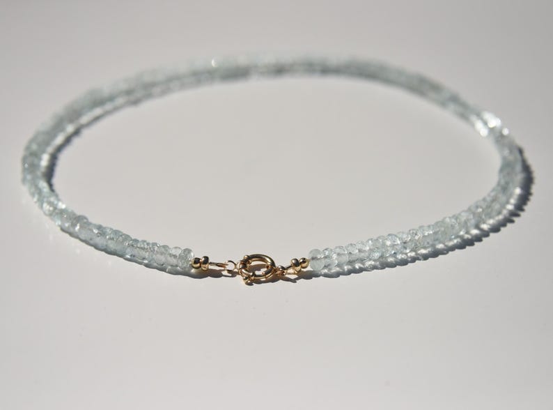 May include: A delicate aquamarine necklace with a gold clasp. The necklace is made of small, light blue gemstones, strung together to form a double strand. The clasp is a small, round design with a gold finish.