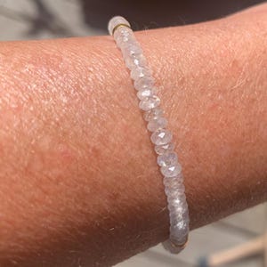 May include: A delicate bracelet featuring small, faceted, clear beads strung together. The bracelet is accented with gold-colored metal elements. The beads appear to be made of a translucent material, reflecting light.