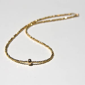 May include: A delicate gold necklace with a small cylindrical pendant. The necklace is made of tiny, closely spaced beads, creating a shimmering effect. The pendant is centered on the chain, adding a touch of elegance to the minimalist design.