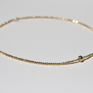 May include: Delicate gold necklace featuring small, evenly spaced beads. The necklace has a simple, elegant design with a single, larger bead near the clasp. The jewelry is displayed on a white surface, highlighting its golden color.