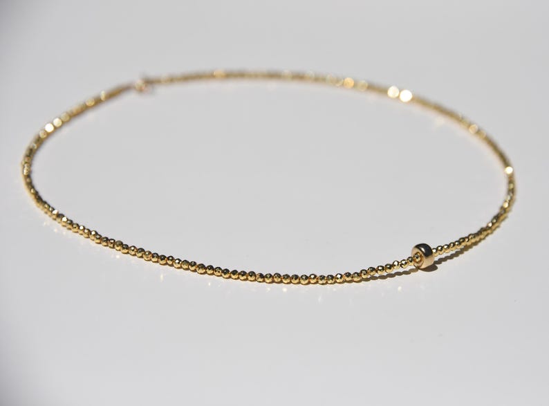May include: Delicate gold necklace featuring small, evenly spaced beads. The necklace has a simple, elegant design with a single, larger bead near the clasp. The jewelry is displayed on a white surface, highlighting its golden color.