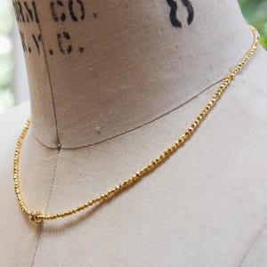 May include: A delicate gold necklace with small, faceted beads. The necklace is displayed on a mannequin, showcasing its length and design. The gold color provides a warm, elegant look, suitable for everyday wear or special occasions.