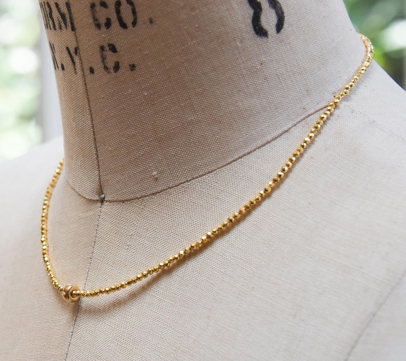 May include: A delicate gold necklace with small, faceted beads. The necklace is displayed on a mannequin, showcasing its length and design. The gold color provides a warm, elegant look, suitable for everyday wear or special occasions.