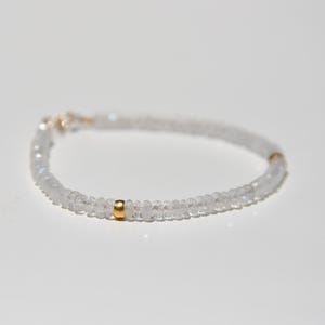 May include: Delicate bracelet featuring small, clear, round beads. The bracelet has a gold-colored clasp and a gold-colored accent bead. The beads are strung together to form a flexible band, creating a minimalist and elegant design.
