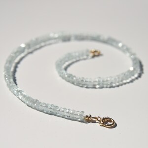 May include: A light blue aquamarine gemstone necklace with a gold clasp. The necklace is made of small, faceted beads. The clasp is a small, gold-coloured hook and eye closure. The necklace is displayed on a white surface.