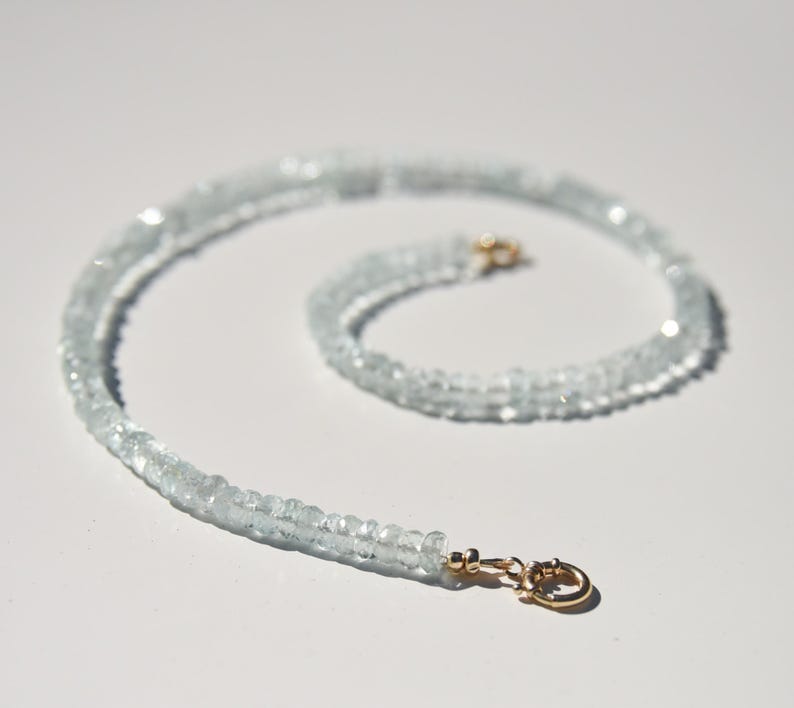 May include: A light blue aquamarine gemstone necklace with a gold clasp. The necklace is made of small, faceted beads. The clasp is a small, gold-coloured hook and eye closure. The necklace is displayed on a white surface.