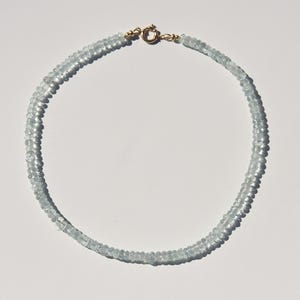 May include: A delicate aquamarine gemstone necklace. The necklace features small, light blue aquamarine beads strung together. The clasp is gold-coloured. The necklace is displayed on a white surface.