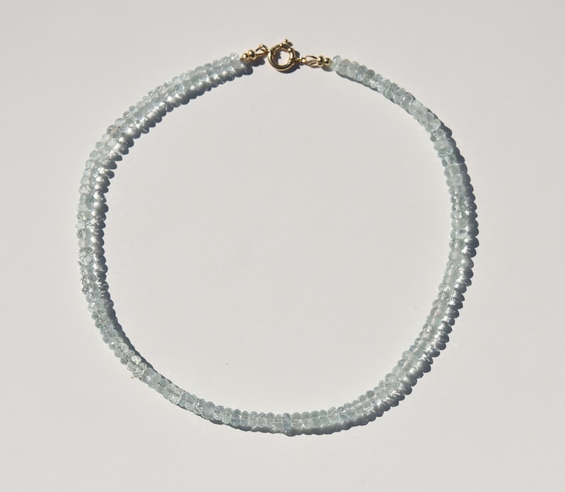 May include: A delicate aquamarine gemstone necklace. The necklace features small, light blue aquamarine beads strung together. The clasp is gold-coloured. The necklace is displayed on a white surface.